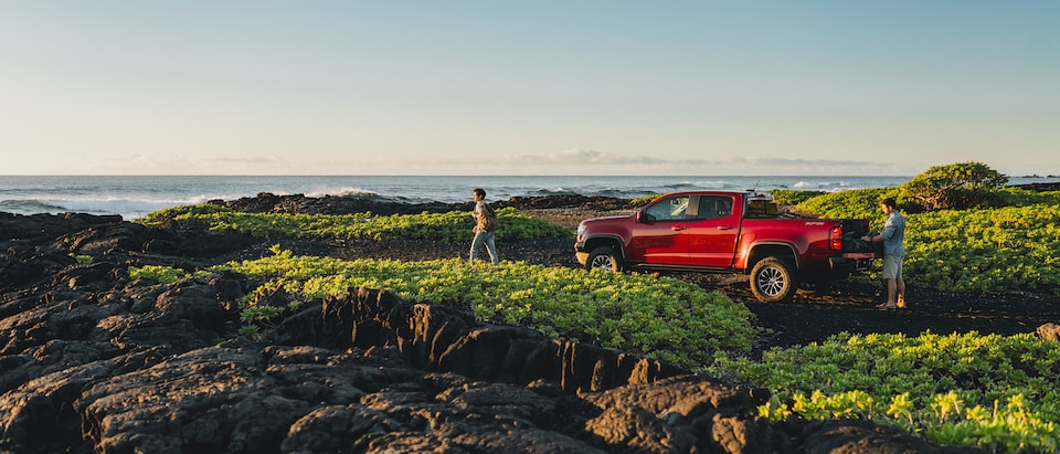 Two people stand near a red Chevrolet Colorado ZR2 parked in a rocky area with low brush next to the ocean in Hawaii.