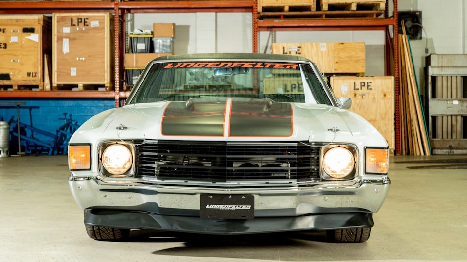 Seen from directly in front of the vehicle, a 1972 El Camino, gray with black racing stripes on the hood, sits in a warehouse building. A bright red graphic across the top of the windshield reads LINGENFELTER.