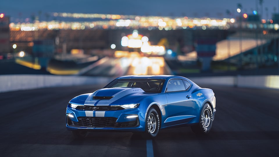 A blue COPO Camaro sits on an empty road with city lights in the background.