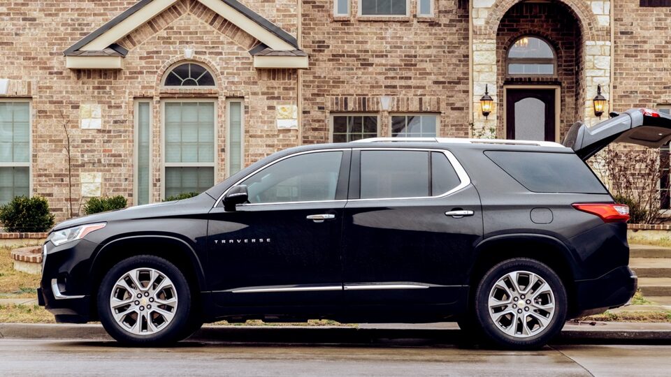 A black Chevy Traverse is parked curbside in front of a brick building with its back hatch open.