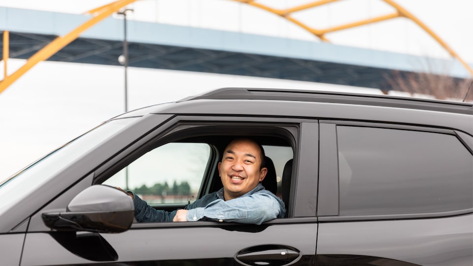 Erik Kennedy smiles as he looks out the open driver's side window of his black 2021 Trailblazer RS, parked below a blue bridge with yellow arch supports.