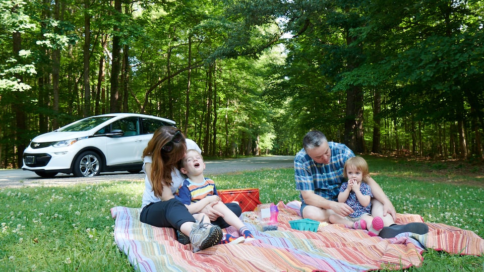 Un hombre y una mujer y dos niños están sentados sobre una manta colorida en el césped disfrutando un picnic, con un Bolt EV en blanco en el fondo.