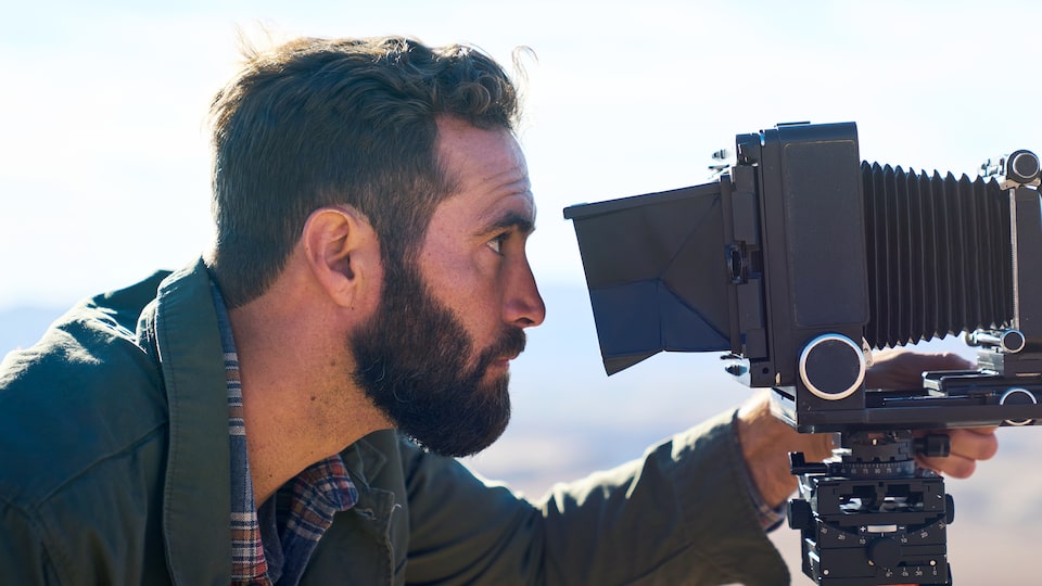A man with a beard looks through the lens of a large-format camera in front of an out-of-focus background of the high Arizona desert.