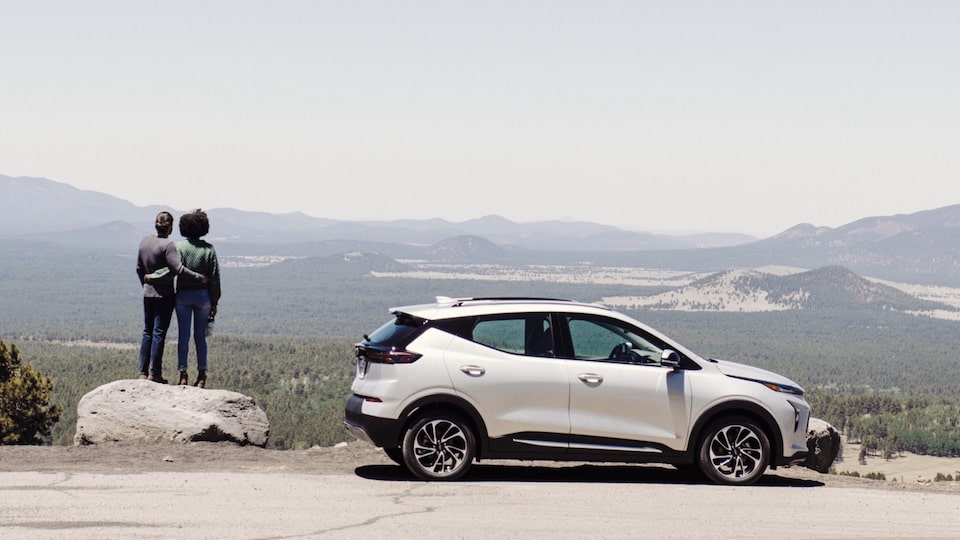 A white Chevy Bolt EUV sits in a pullout on a road in northern Arizona with a mountain landscape in the background. Two people stand on a large boulder next to the car.
