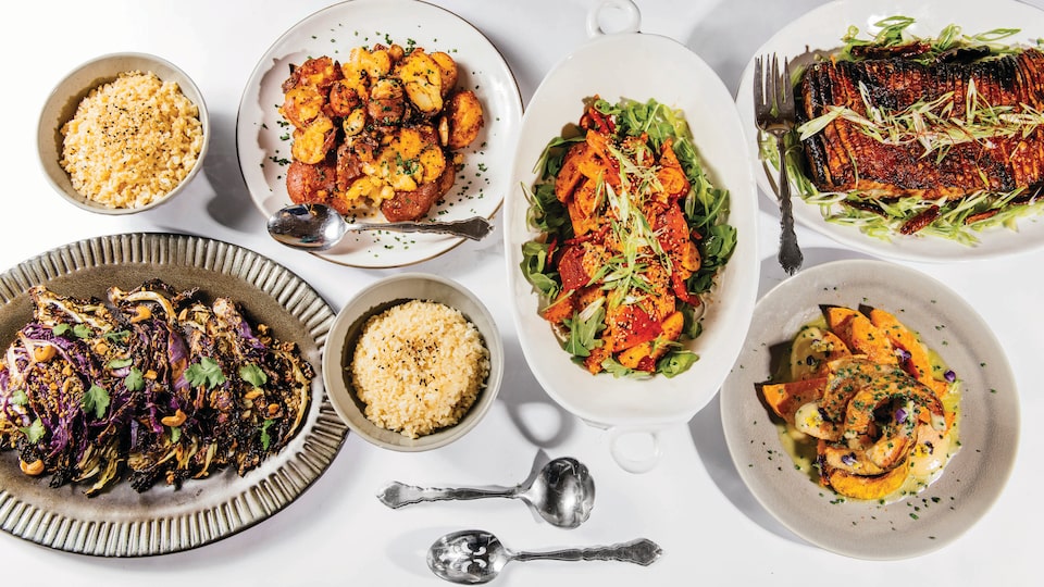 Looking down from above, several plates and platters filled with artfully arranged, colorful food sit on a white tabletop.