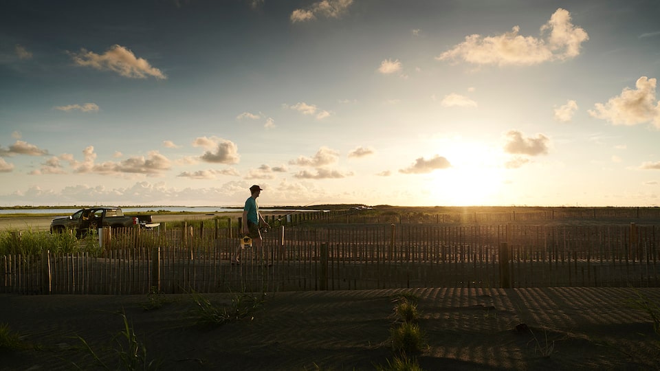 A man with a fishing pole and tackle box walks along a boardwalk at dawn next to a saltwater marsh, with a Chevy Silverado in the background.