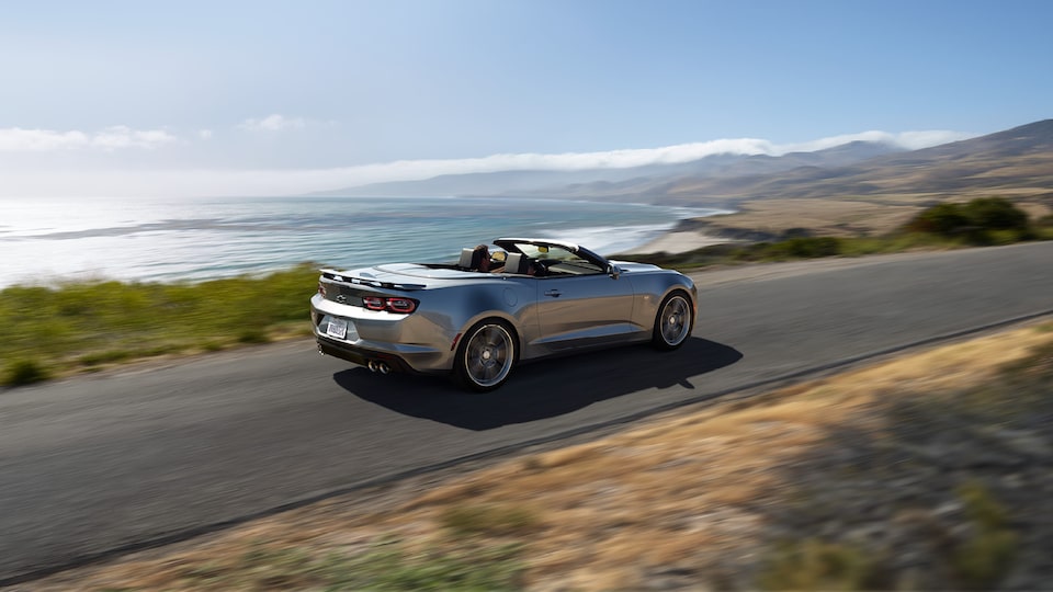 A Silver Ice Metallic Camaro Convertible driving down a road high above the ocean with mountains in the distance.