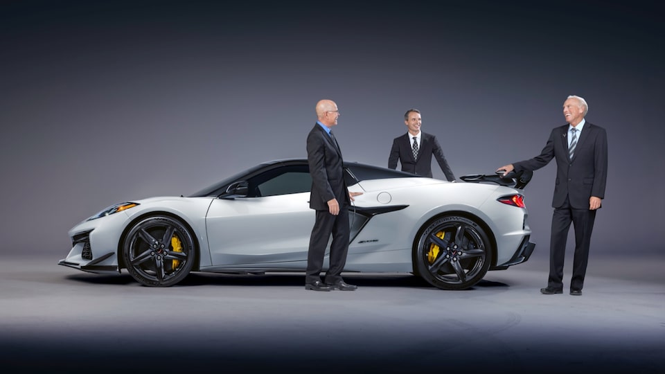 Executive chief engineer Tadge Juechter, global chief engineer Jordan Lee, and vehicle performance manager Alexander MacDonald, all wearing black suits, stand around a white Corvette Z06 with black trim that is sitting on a black floor in front of a black background.