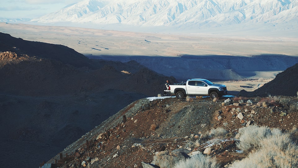 A silver Chevy Colorado ZR2 Bison pickup truck sits on a high ledge atop a hill in the California mountains, with the desert and mountains in the background.