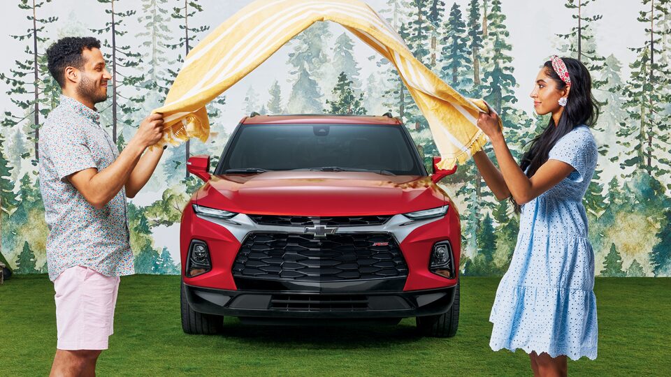 Two people face each other in front of a Cherry Red Tintcoat Chevy Blazer RS as they shake out a yellow striped picnic blanket between them.