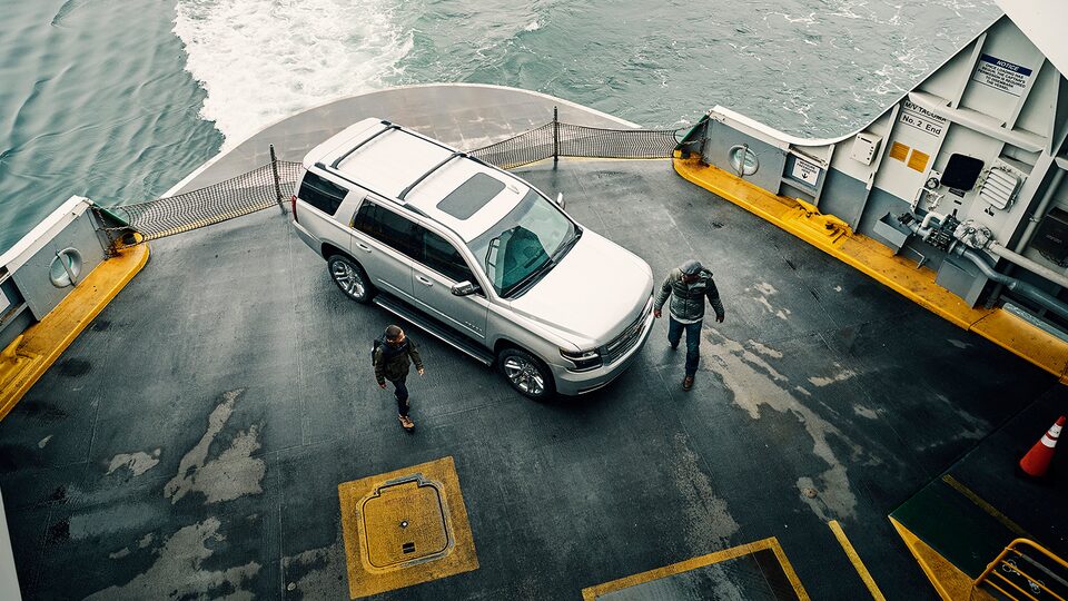 A son and father walking past a Chevy Tahoe SUV on the deck of a ferry that’s underway.