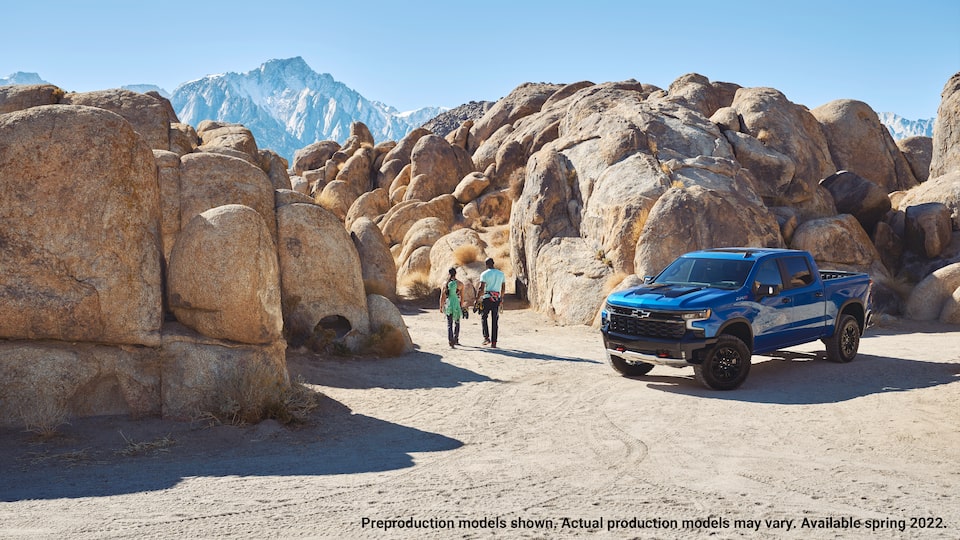 A blue Silverado ZR2 pickup sits on a flat gravel area with high, rounded boulders behind it and mountains in the background. Two people carrying climbing gear walk away from the truck toward the boulders.