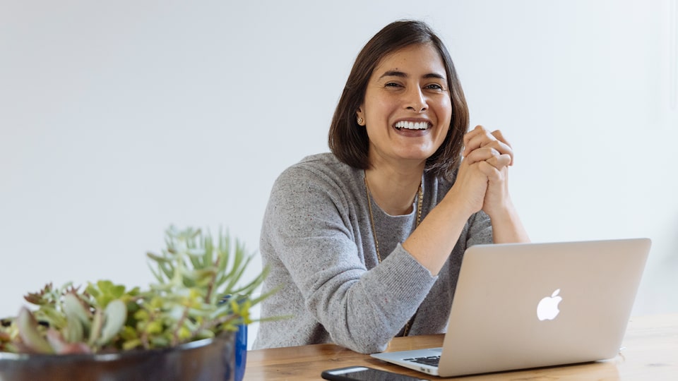 A woman sits at a table smiling with an open laptop and cellphone in front of her.