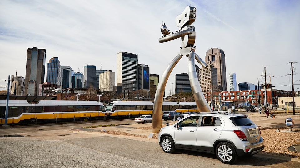 Una Chevrolet Trax color plata en la calle al lado de una escultura moderna con el horizonte de Dallas en el fondo.