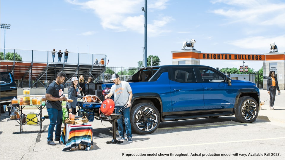 Un grupo de personas reunidas cerca de la puerta trasera de una Silverado EV con una parrilla portátil y una mesa portátil con comida y bebida, además de otros artículos en la puerta trasera de la Silverado.