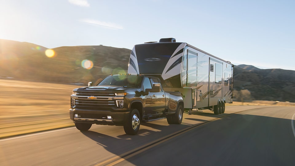 A 2020 black Silverado HD pickup hauls a large travel trailer down a two-lane road with hills in the background.