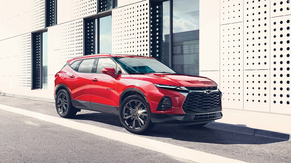 The front and passenger side of a red Blazer RS SUV parked next to a contemporary-style building