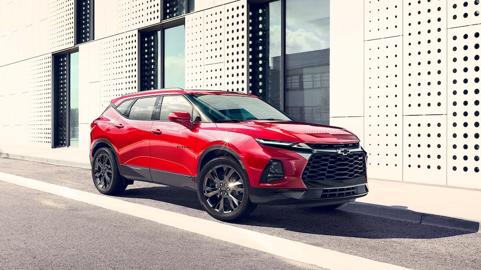 The front and passenger side of a red Blazer RS SUV parked next to a contemporary-style building.