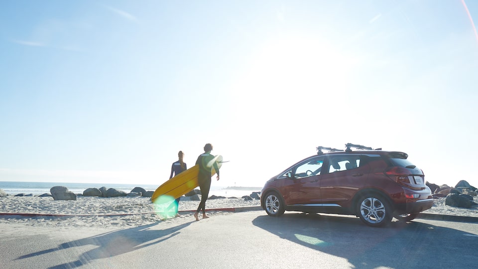 Two people in wetsuits walk from a parking lot out onto an ocean beach carrying a surfboard. A red Chevrolet Bolt EV sits in the parking lot.
