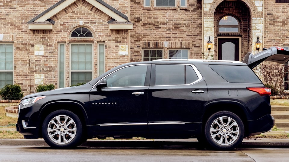 A black Chevy Traverse is parked curbside in front of a brick building with its back hatch open.