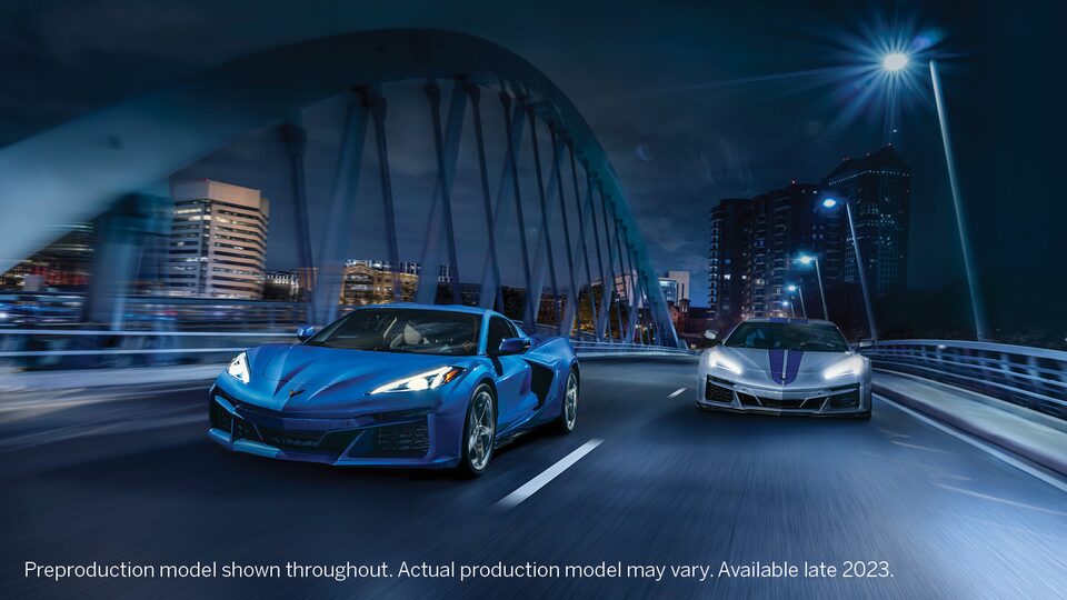 A blue Corvette E-Ray drives across a bridge at night next to a gray Corvette E-Ray with blue racing stripes.