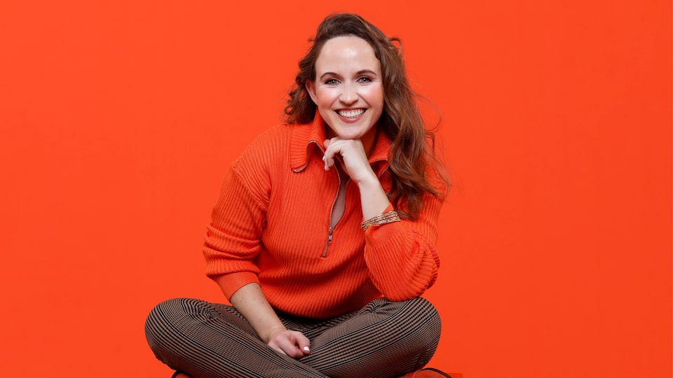 Katie Sloman sits cross-legged in front of a bright red background and smiles at the camera.