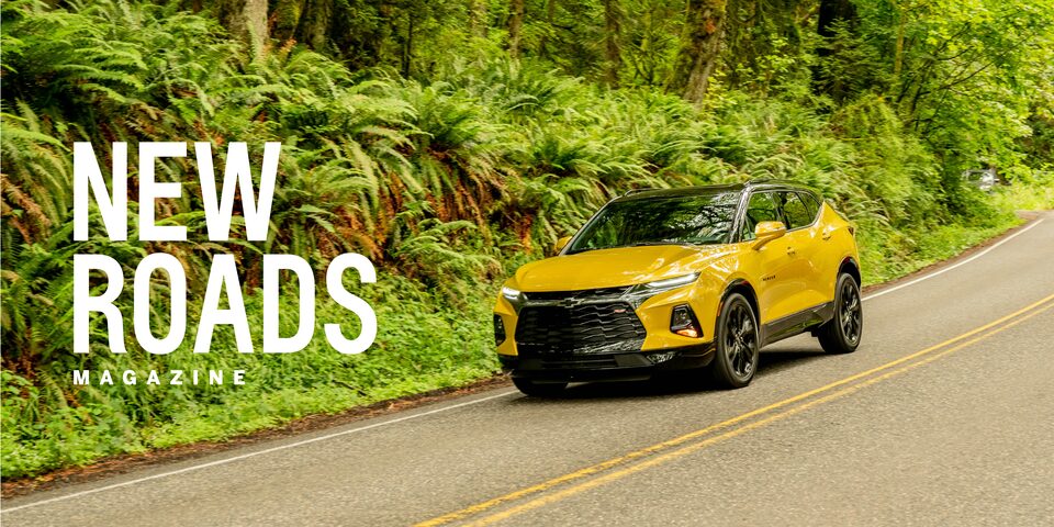 A bright yellow Blazer drives down a two-lane road that is lined with trees. The words “New Roads Magazine” appear over the image.