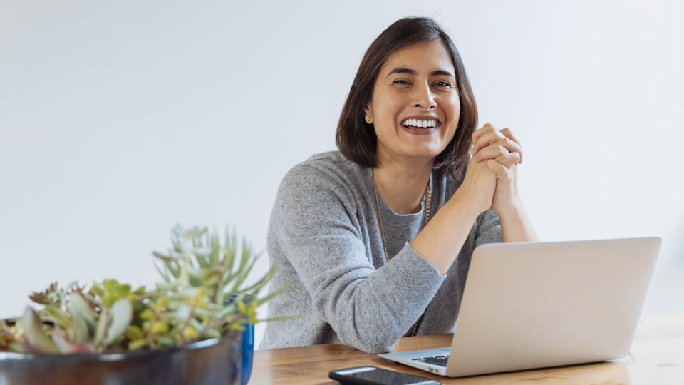 A woman sits at a table smiling with an open laptop and cellphone in front of her.