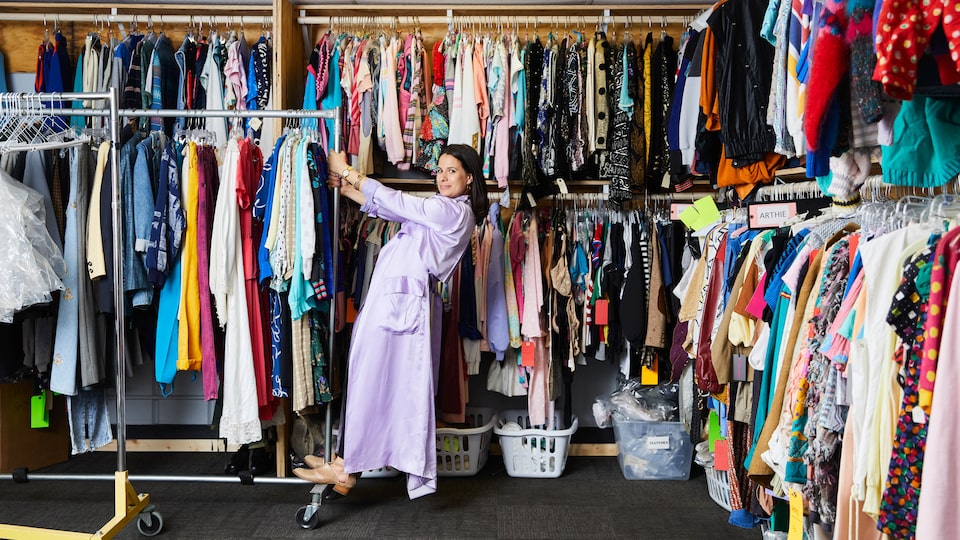 Beth Morgan stands on a portableclothes rack filled with costumes in front of wall racks filled end-to-end with costumes.