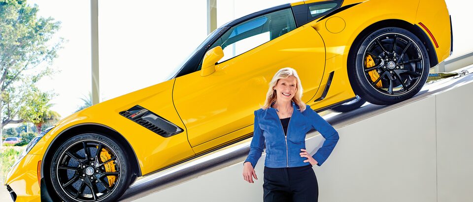 Hertz CEO Kathy Marinello with a yellow Chevrolet Corvette displayed on a ramp behind her.
