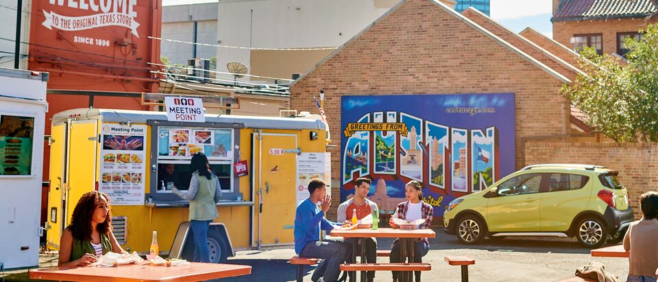 Several people eating at tables at the University Co-op food truck area in Austin, Texas, with a yellow 2018 Chevy Spark in the background.