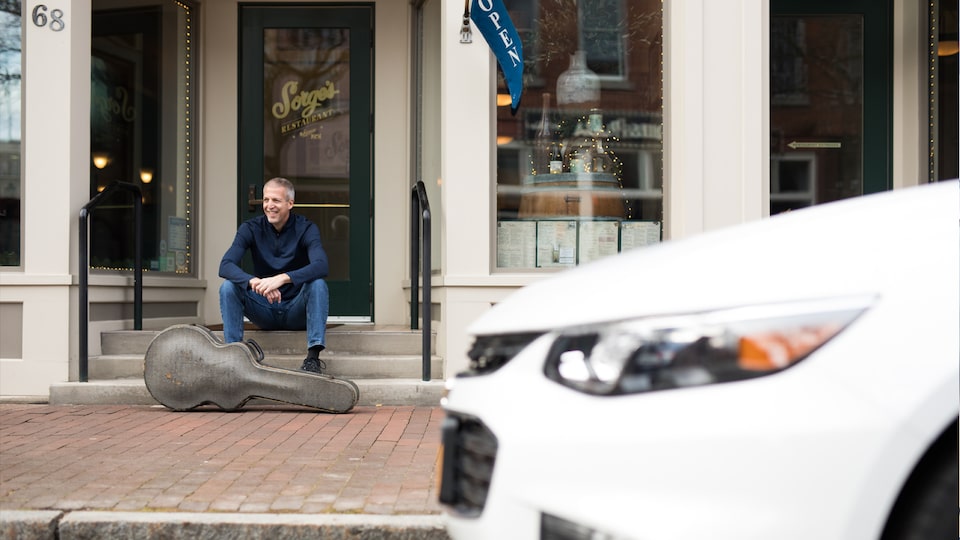 Mike Lucey sitting on a bench with his guitar case, with his Chevrolet Malibu in the foreground.