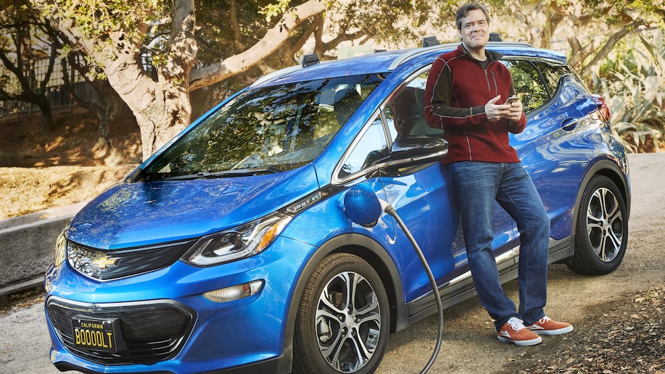 Ethan Stock standing next to his blue 2017 Chevy Bolt EV, which is plugged in to charge.