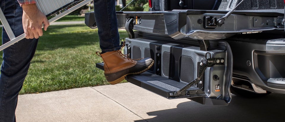 A person wearing work boots and carrying a ladder has one foot up on the Silverado Multi-Flex tailgate configured as a step.