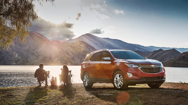 A couple sits in lawn chairs on the shore of a lake next to a Chevrolet Equinox.