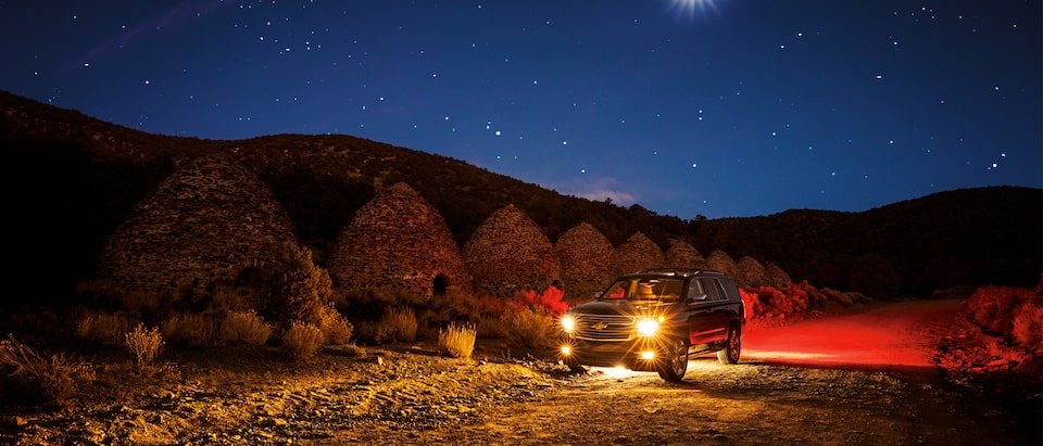 Una Chevy Tahoe 2018 por la noche en el desierto, bajo la luz de las estrellas y la luna, y un paisaje iluminado por los faros delanteros y traseros de la Tahoe.