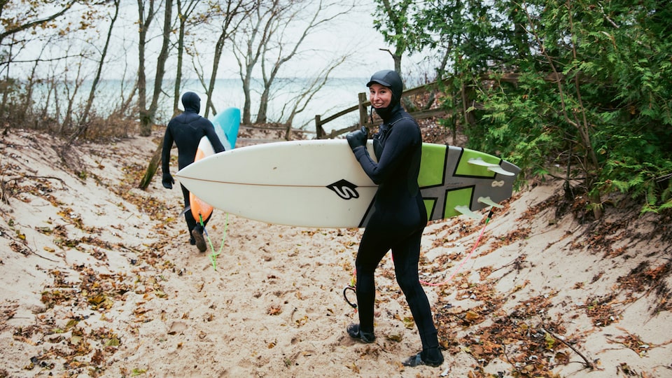 Two surfers in wetsuits walking down a sandy path toward Lake Michigan.