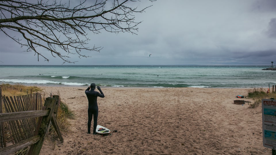 A surfer in a wetsuit stands on a Lake Michigan beach looking out at the water and gray sky.