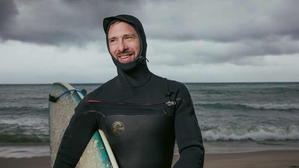 A surfer in full gear holds his surfboard on a Lake Michigan beach with waves crashing behind him.