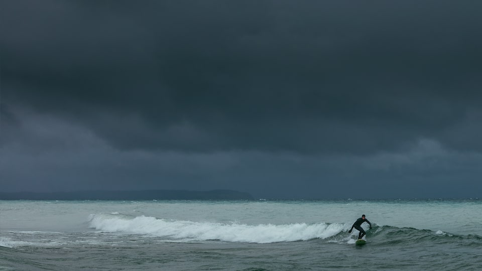 A surfer rides a wave in on dark Lake Michigan water with an ominous gray sky behind him.