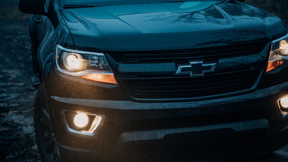 The front end of a black Chevy Colorado Z71 Crew Cab Midnight Edition in semi-darkness with headlights and foglamps glowing.