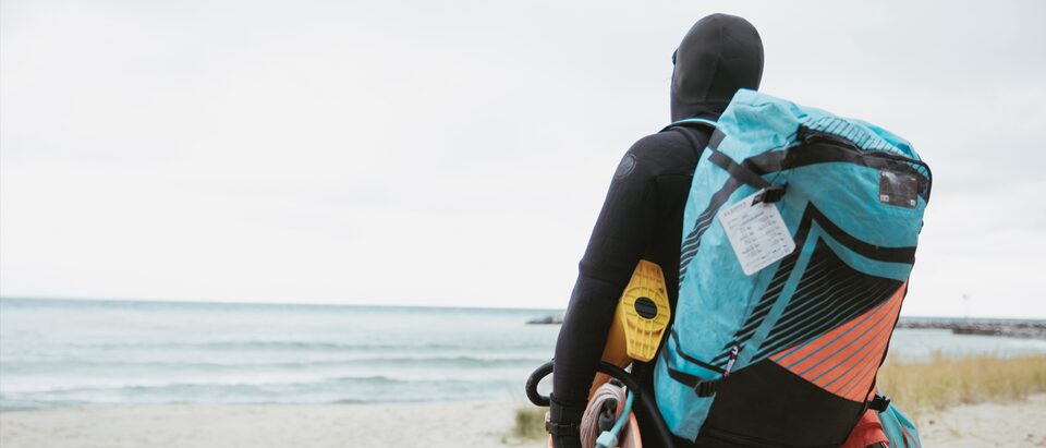 Persona con traje de neopreno caminando por la playa hacia el Lago Michigan con la mochila repleta y transportando su equipaje.