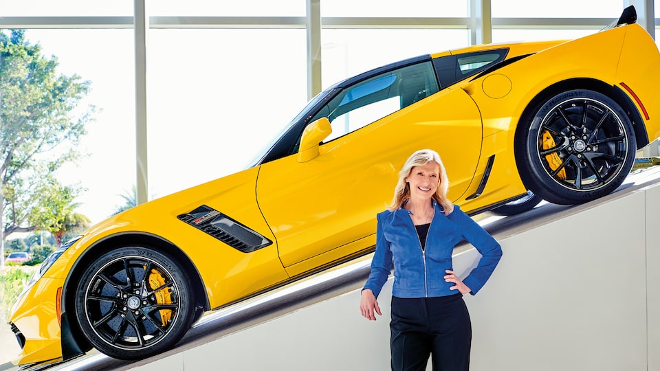 Hertz CEO Kathy Marinello with a yellow Chevrolet Corvette displayed on a ramp behind her.