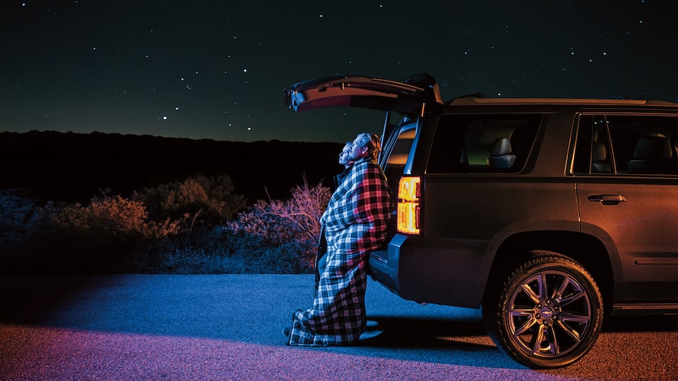 Two people standing under the open back hatch of a 2018 Chevy Tahoe, wrapped in blankets and gazing up at the star-filled sky.