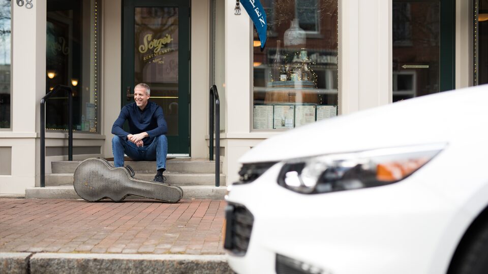 Mike Lucey sitting on a bench with his guitar case, with his Chevrolet Malibu in the foreground.