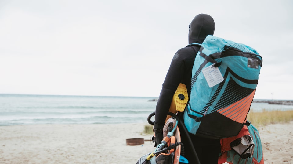 Persona con traje de neopreno caminando por la playa hacia el Lago Michigan con la mochila repleta y transportando su equipaje.