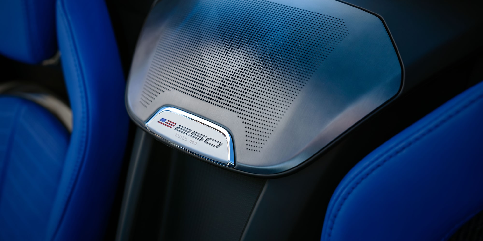 A Close-Up View Of A Chevrolet Interior Showcasing A Silver America250 Badge Integrated Into A Textured Speaker Cover Between Blue Leather Sport Seats.
