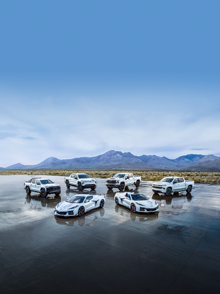 White Chevy Trucks and Corvettes Parked in a Field with Mountains in the Background.