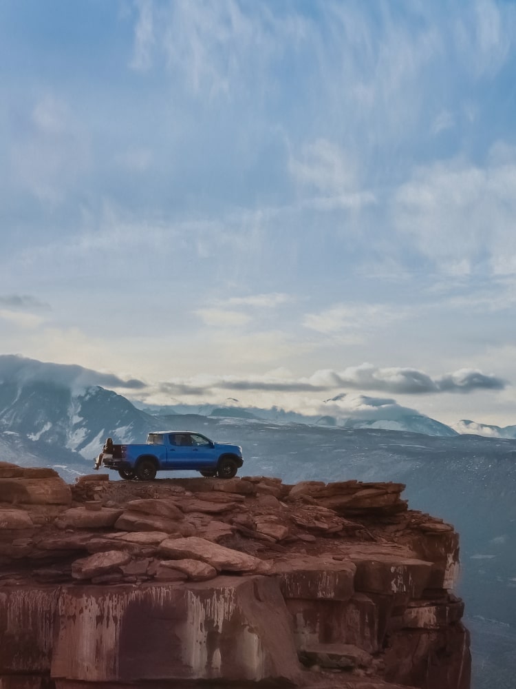Una camioneta Chevrolet en Azul estacionada al borde de un enorme acantilado rocoso, con una cadena montañosa de fondo, al atardecer.