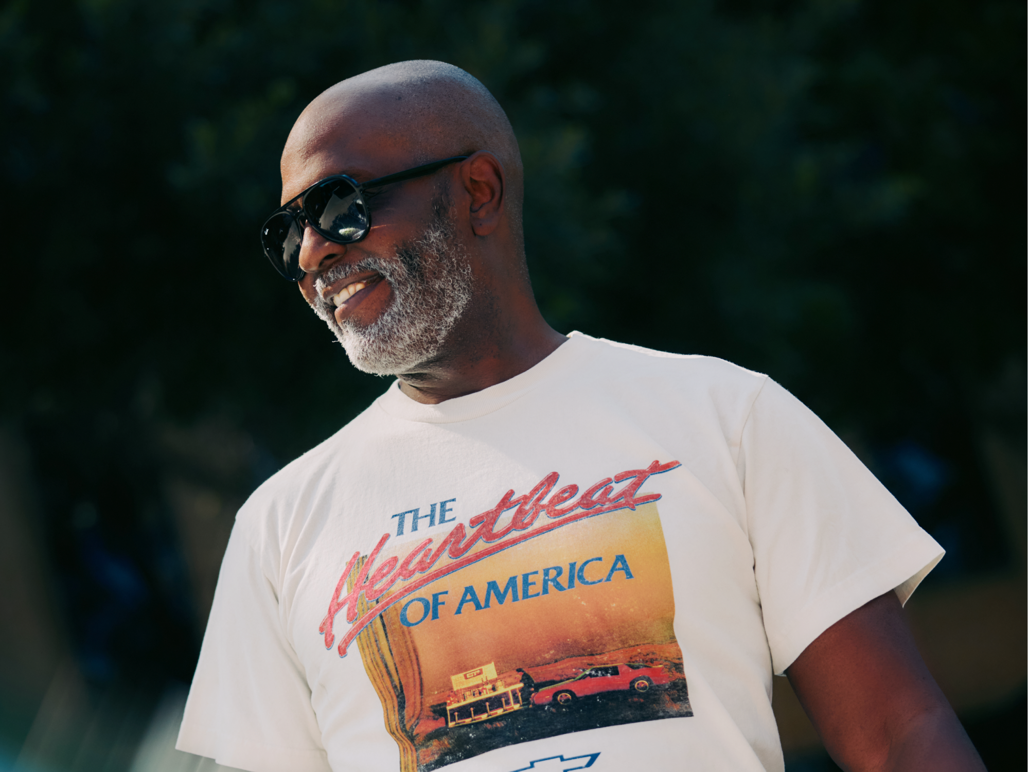 Un hombre sonriendo con gafas de sol puestas y una camisa que dice "The Heartbeat of America"